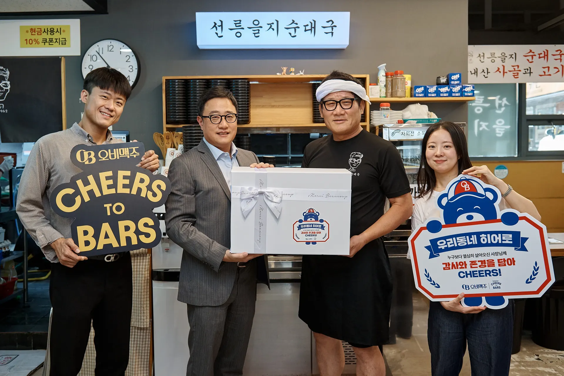 오비맥주 관계자와 소상공인 점주가 선물 박스를 함께 들고, ‘CHEERS TO BARS’와 ‘우리동네 히어로’ 문구의 피켓을 들며 소상공인 지원 캠페인을 알리는 기념촬영 모습