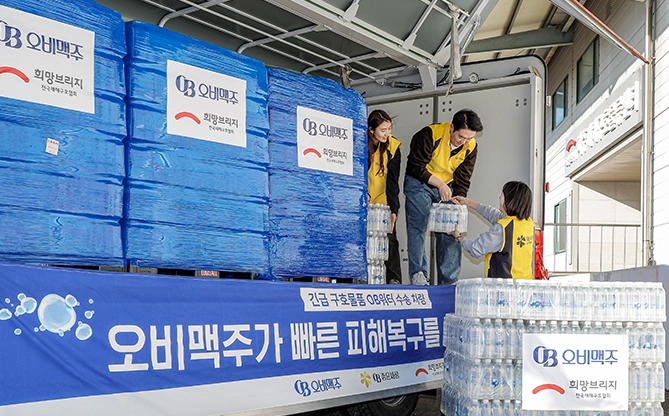 오비맥주와 희망브리지 로고가 붙은 구호 물품 트럭에서 임직원들이 구호용 생수(OB워터)를 옮기고, 대량의 생수가 현장에 적재된 재해구호 지원 모습이 있습니다.
